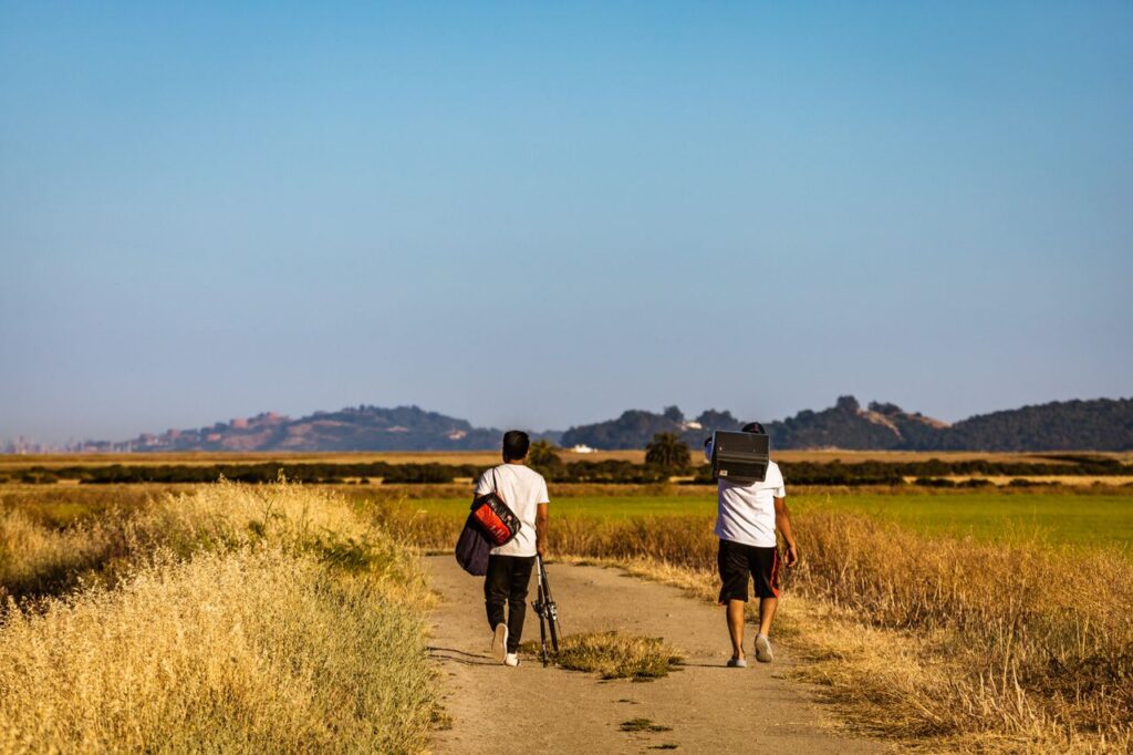 Two men walking on a rural trail with a scenic hillside view under a clear blue sky.